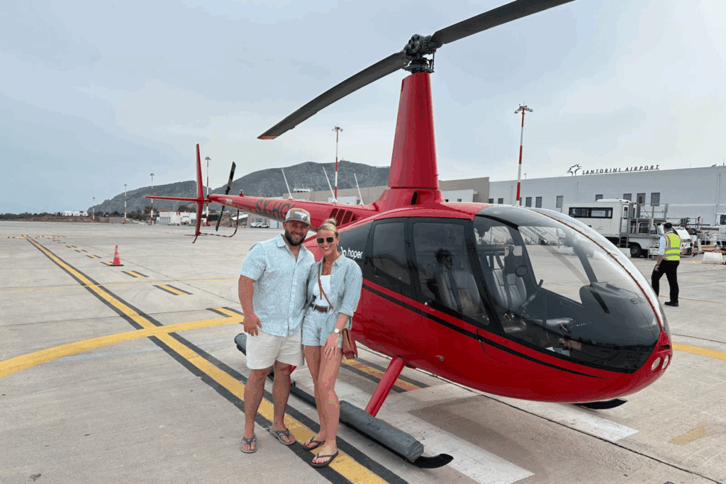 Two people stand proudly in front of a red helicopter at Santorini Airport, ready for a private sightseeing and wine tour.
