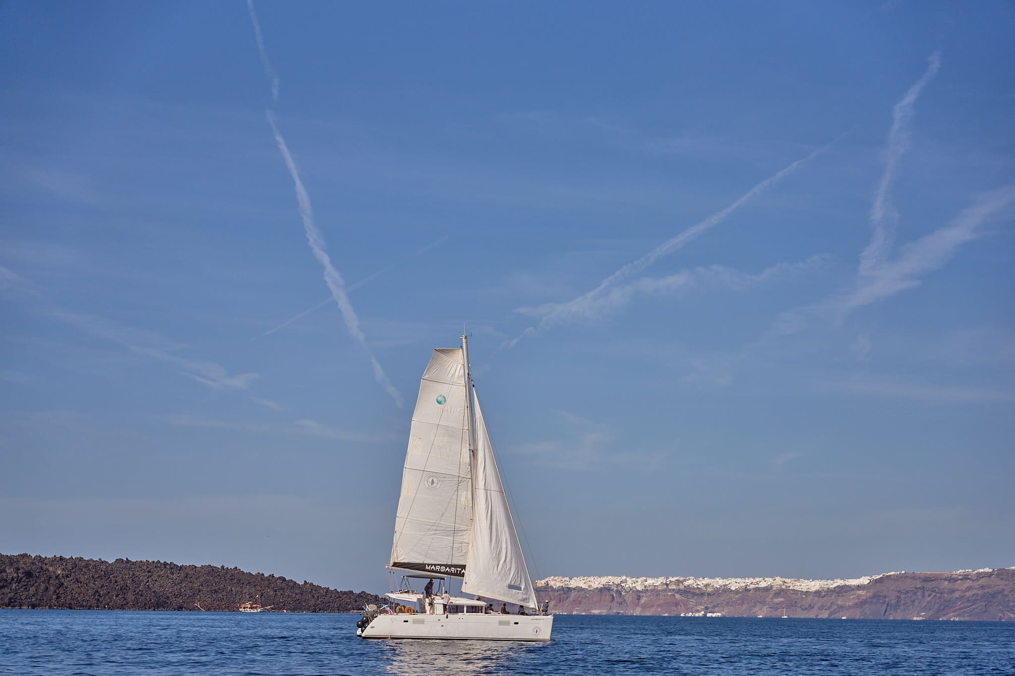 Sailing catamaran cruising near the coastline of Santorini with rugged cliffs and clear blue waters in the background.