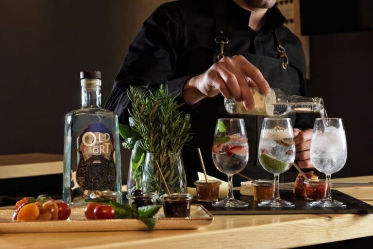 A person in a black apron pours from a bottle into gin-filled glasses, surrounded by garnishes and snacks.
