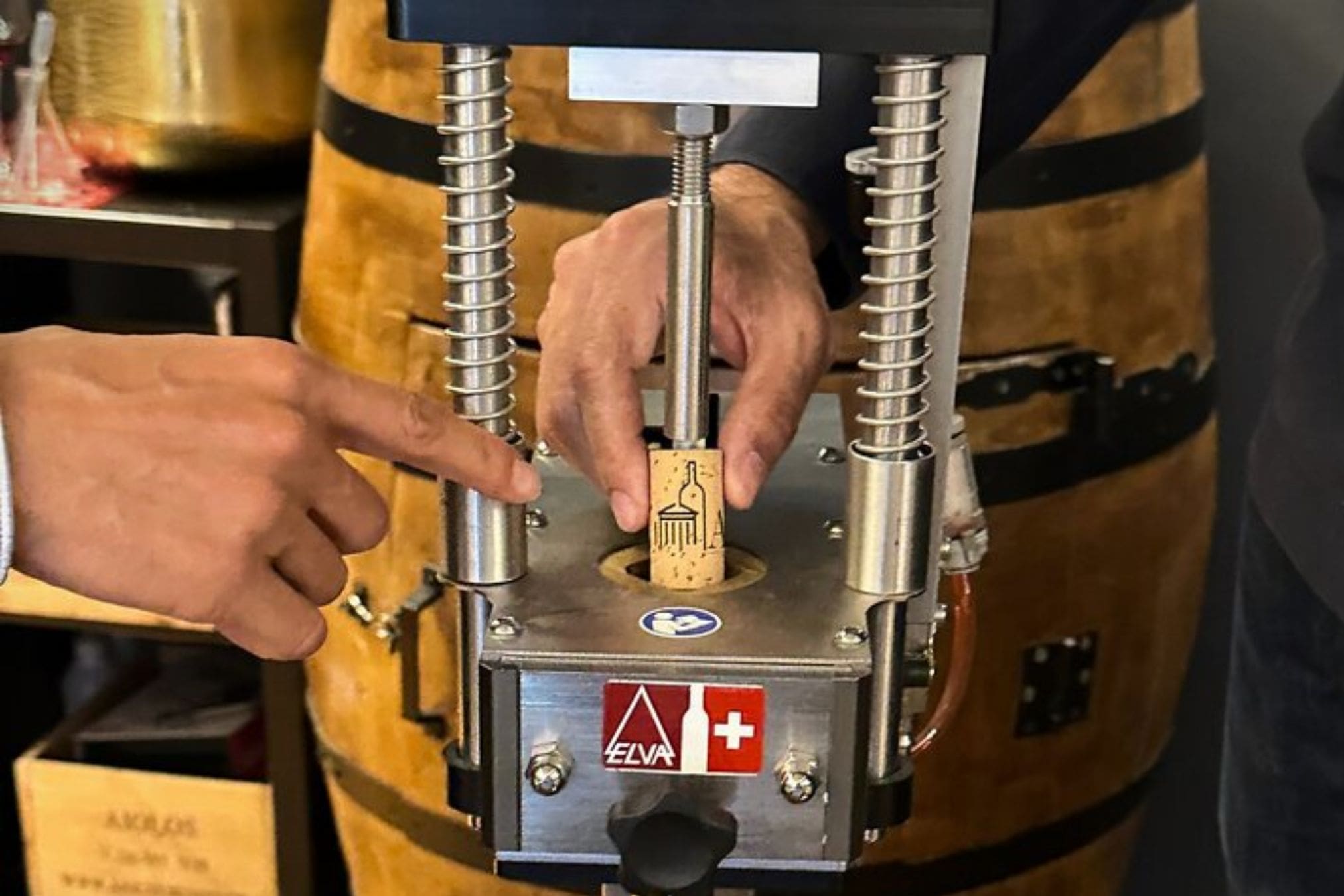 A person uses a corking machine to insert a cork into a wine bottle, with a wine barrel in the background.