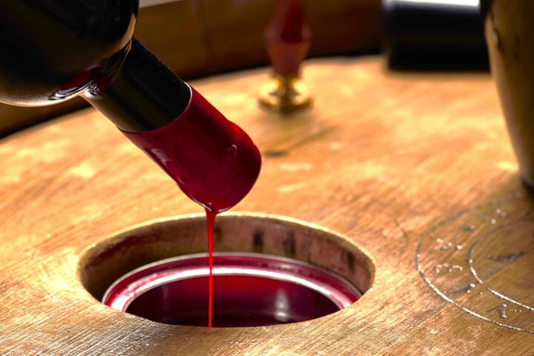 Molten red wax dripping from a wine bottle neck into a round opening on a wooden surface, likely during a bottle sealing process.