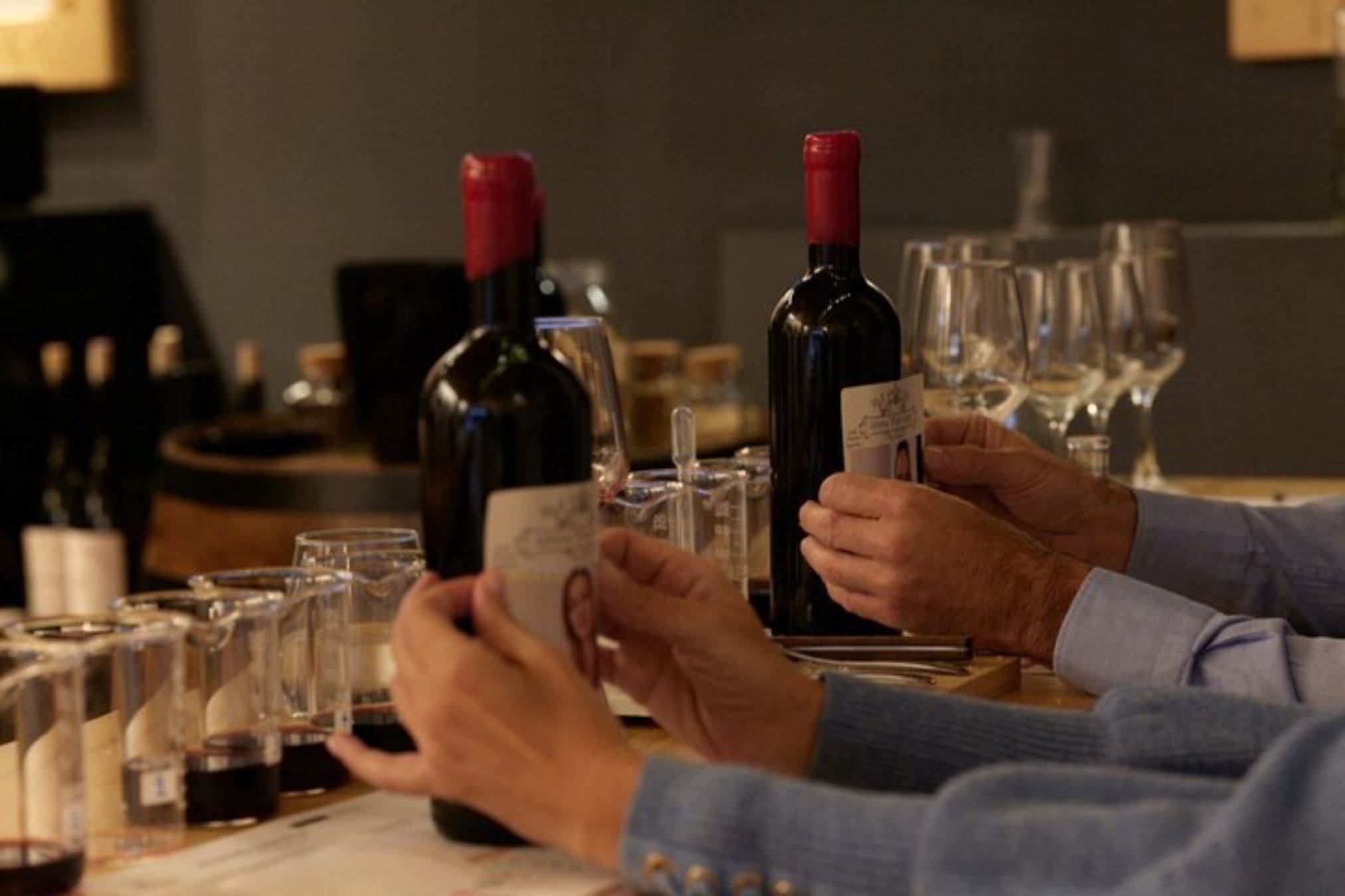 Two people examine wine bottles during a winemaking workshop, with rows of tasting glasses and a wine barrel visible in the background.