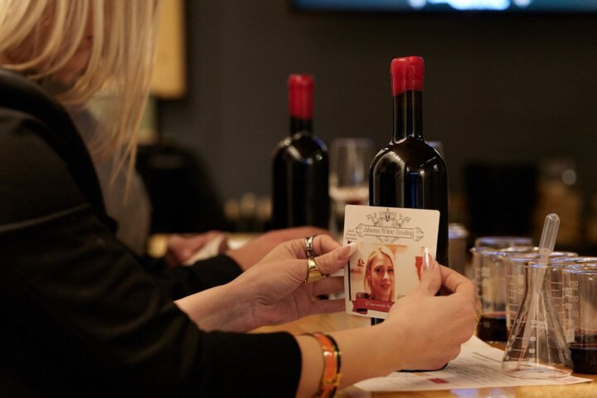A person holds a personalized wine label with a portrait, preparing to affix it to a bottle of wine during an Athens Winemaking Workshop. The image includes wine bottles, glassware, and beakers filled with wine.