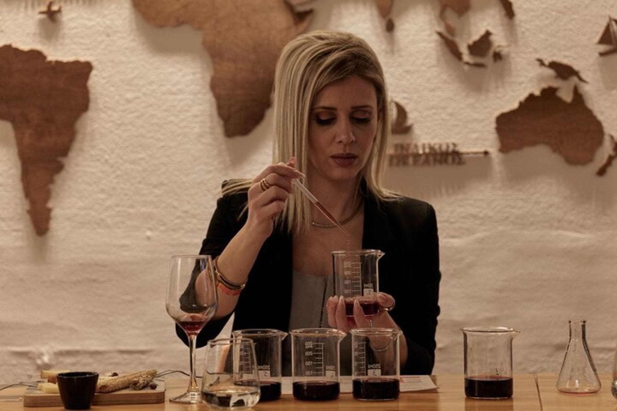 A person uses a pipette to transfer liquid from one beaker to another during a winemaking workshop. A map of the world hangs in the background.