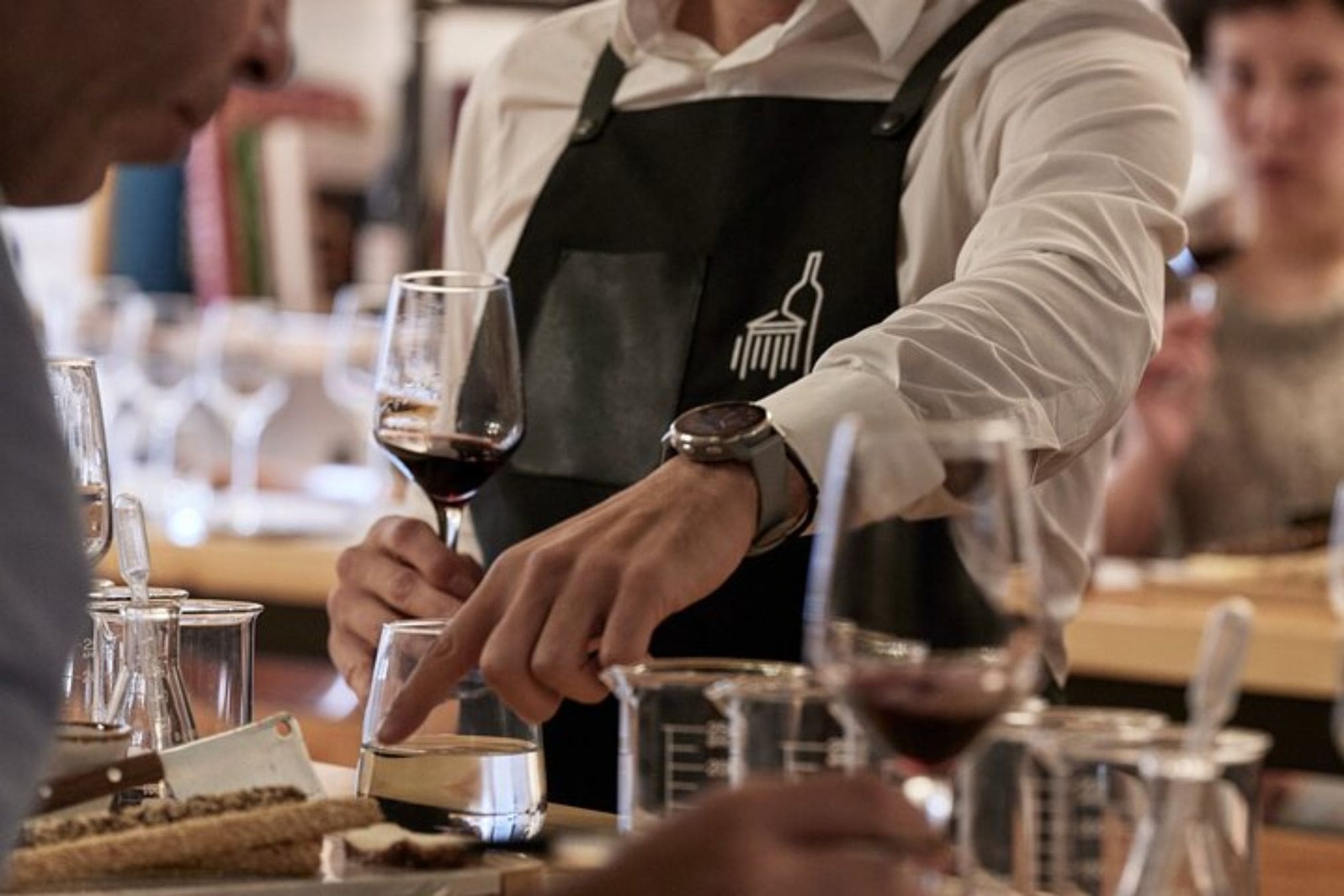 Close-up of a person wearing a black apron with a wine bottle logo, tending to a wine tasting set up with glasses, beakers, and snacks.