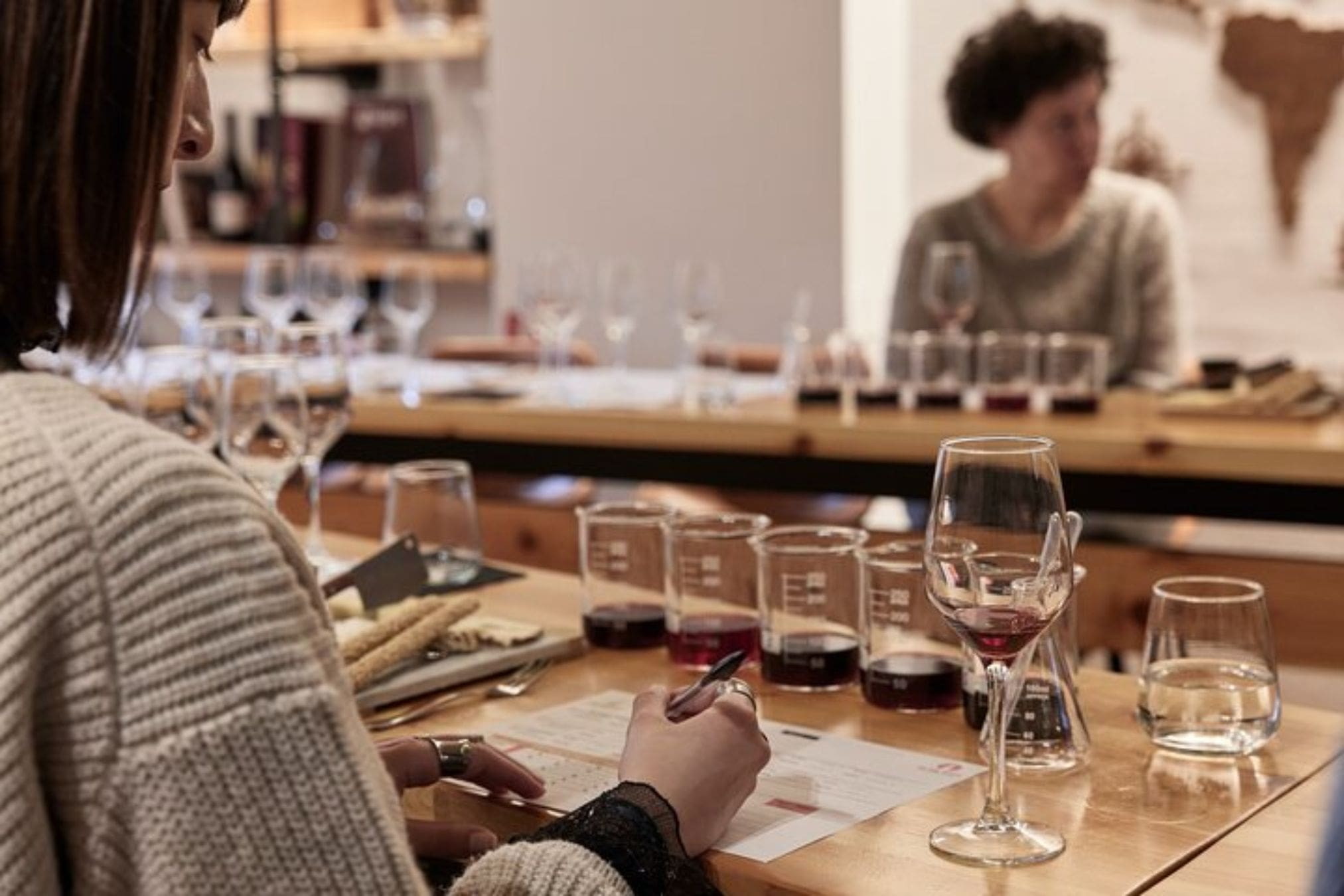 A person is seated at a table, holding a pen and writing on a paper sheet with tasting notes. Several glasses and laboratory beakers containing red wine and water are on the table. In the background, another person is seated, and a world map hangs on the wall.
