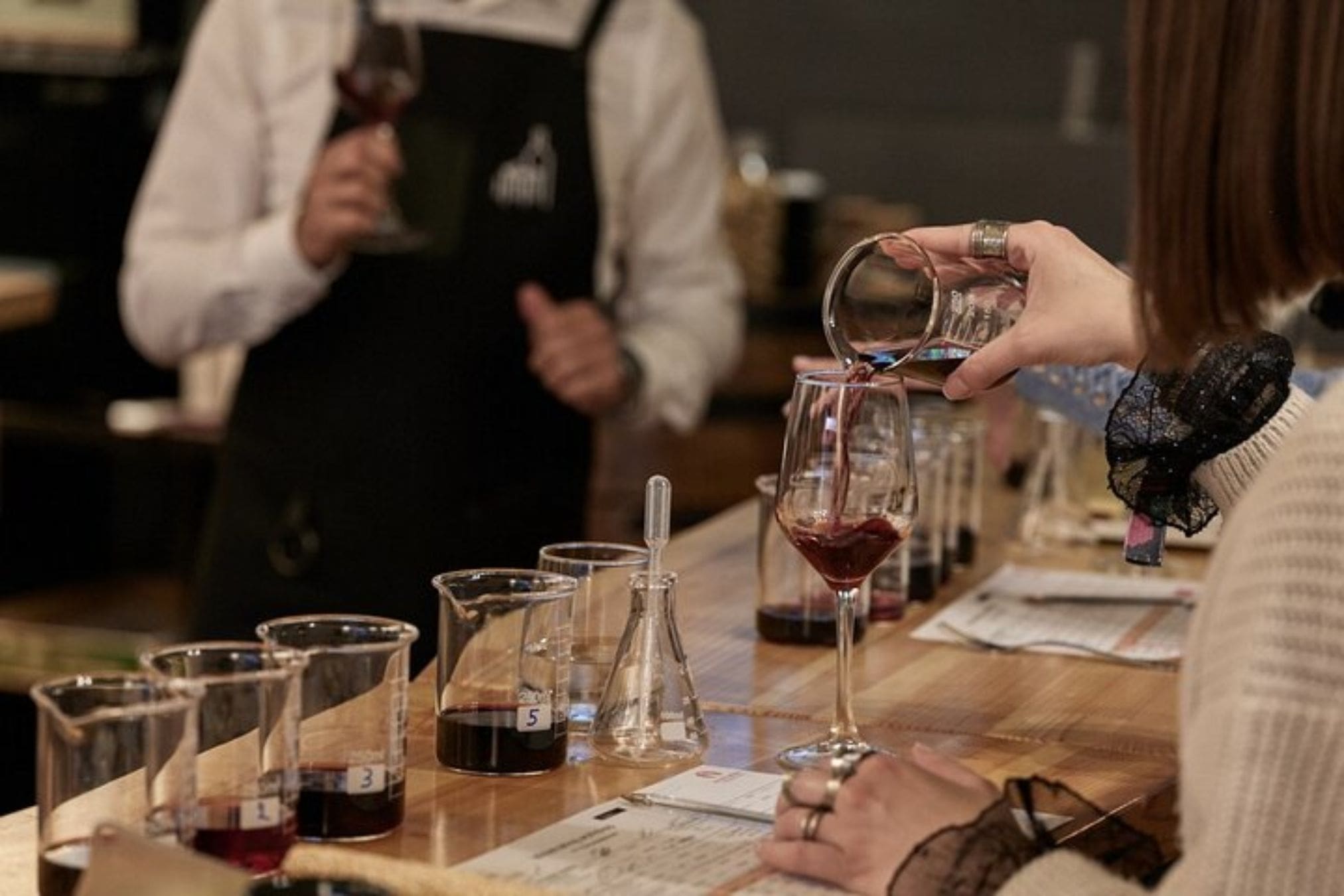A person pours red wine from a beaker into a stemmed wine glass at a winemaking workshop. Various beakers, flasks, and tasting notes are arranged on a wooden countertop. Another person in an apron holds a wine glass in the background.