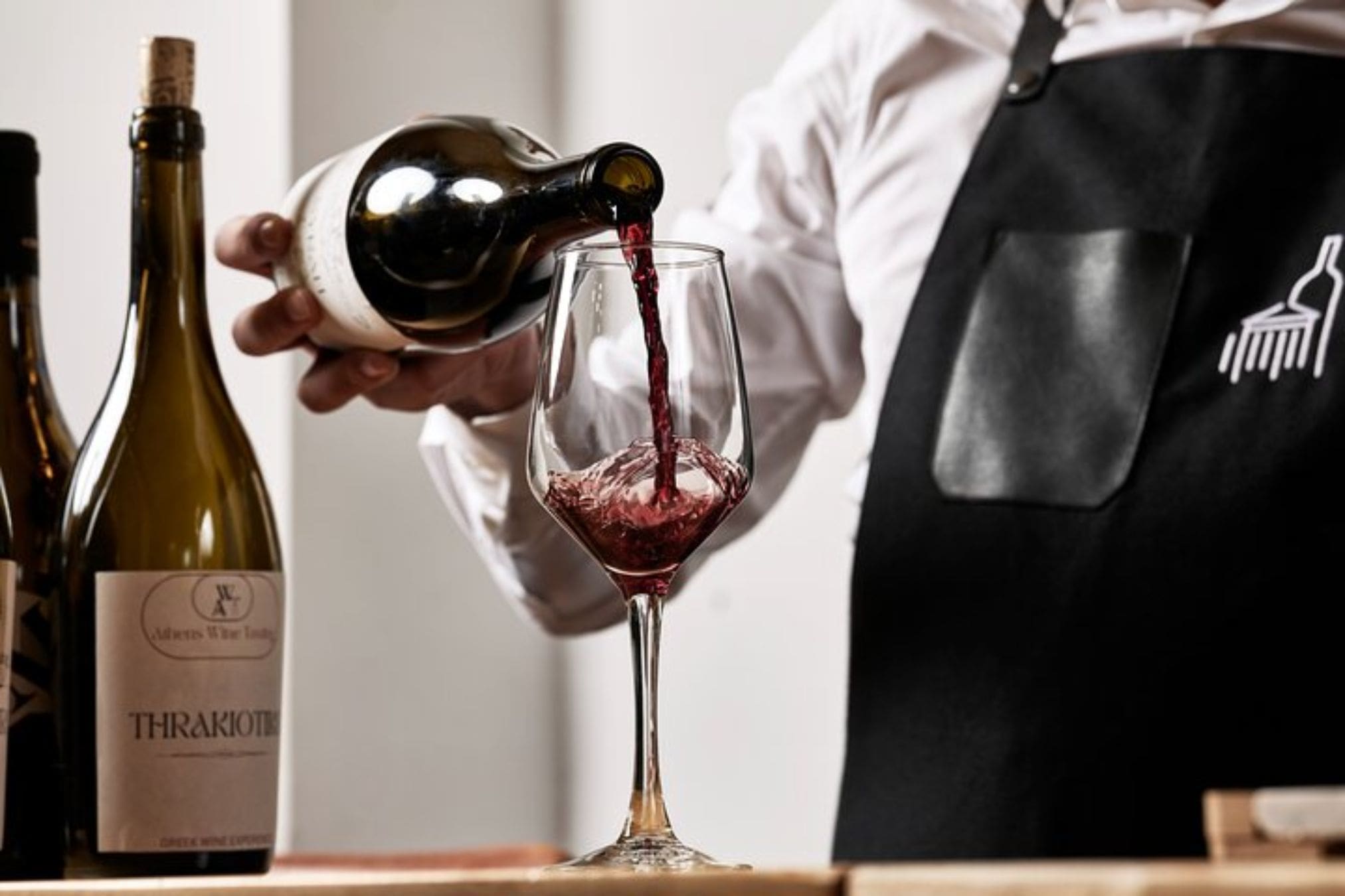 A person in an apron pours red wine into a stemmed glass, with bottles of wine in the background. Title: Athens Winemaking Workshop